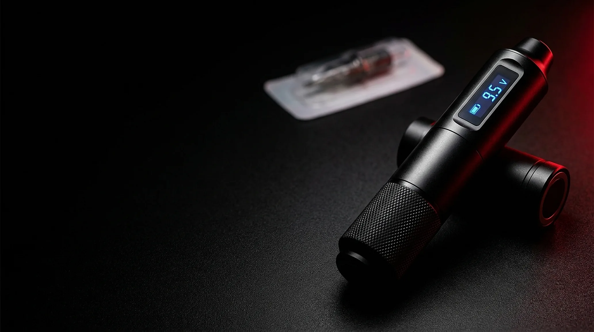 Zwei Tattoo-Maschinen im Pen-Stil auf dunkler Arbeitsflaeche, daneben eine steril verpackte Nadelkartusche
